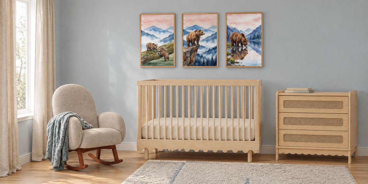 Mountain nursery: three watercolor-style bear and misty mountain prints above a pale birch crib, with a boucle rocker, cane dresser and soft blue-grey walls