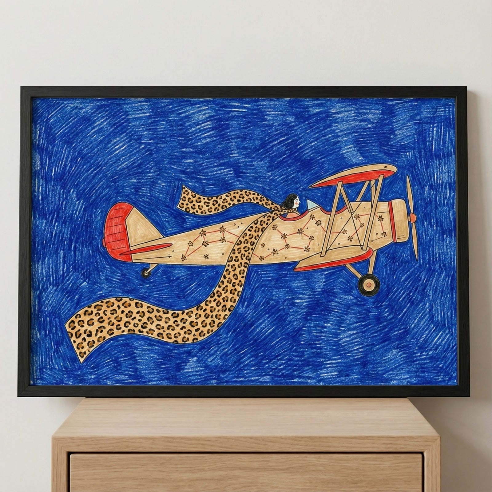Framed biplane wall art featuring a vintage airplane aircraft with star constellations and a pilot wearing a long leopard-print scarf, displayed in a black frame on a light-colored wooden dresser against a plain wall.