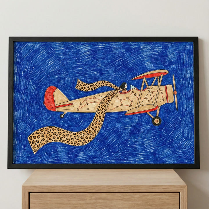 Framed biplane wall art featuring a vintage airplane aircraft with star constellations and a pilot wearing a long leopard-print scarf, displayed in a black frame on a light-colored wooden dresser against a plain wall.