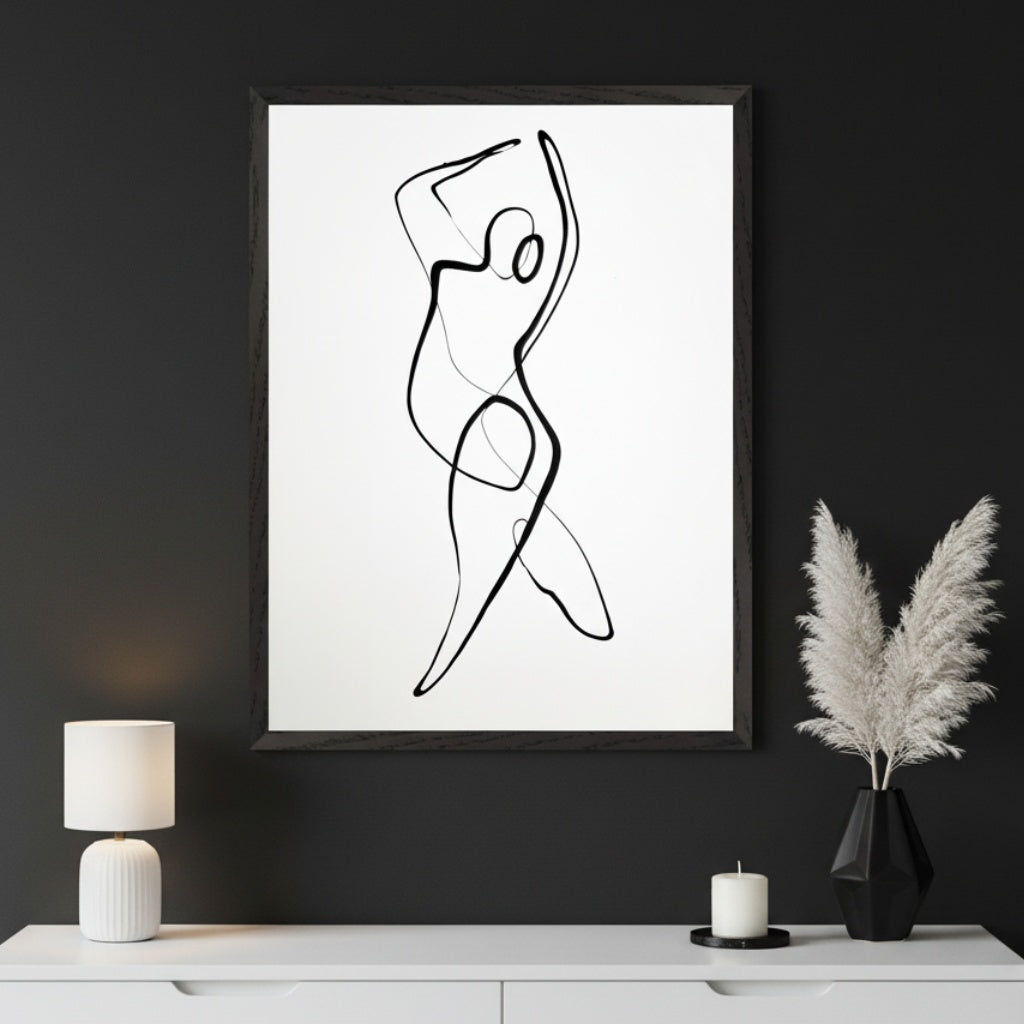 Modern living space with a large black-framed white print depicting a black continuous line drawing of a human figure in motion, centered above a white dresser. A white lamp, a candle, and a black geometric vase with pampas grass are on the dresser. Dark gray wall.