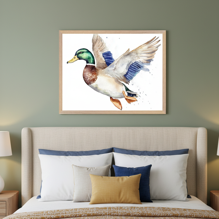 Modern bedroom with a sage green wall and upholstered bed. A framed watercolor painting of a mallard duck in flight, with a green head, brown chest, and blue wing accents, hangs centered above the bed. White, blue, beige, and yellow pillows are on the bed.