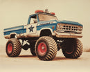 A retro blue and white police truck with oversized monster truck tires sits on a dusty plain. The design includes a star badge logo, red lights on top, and weathered paint, giving it a rugged, law-enforcement-meets-off-road look.