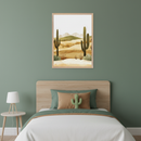 Bedroom with sage green walls, a wooden headboard, and a single bed with white, green, and brown bedding, featuring a cactus pillow. A light wood nightstand with a white lamp sits on the left. A framed desert landscape painting with large saguaro cacti and hills hangs above the bed.