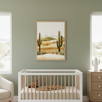 Modern nursery with sage green walls, a white and wood crib, and a light wood dresser. A framed desert landscape illustration with saguaro cacti and hills hangs above the crib.