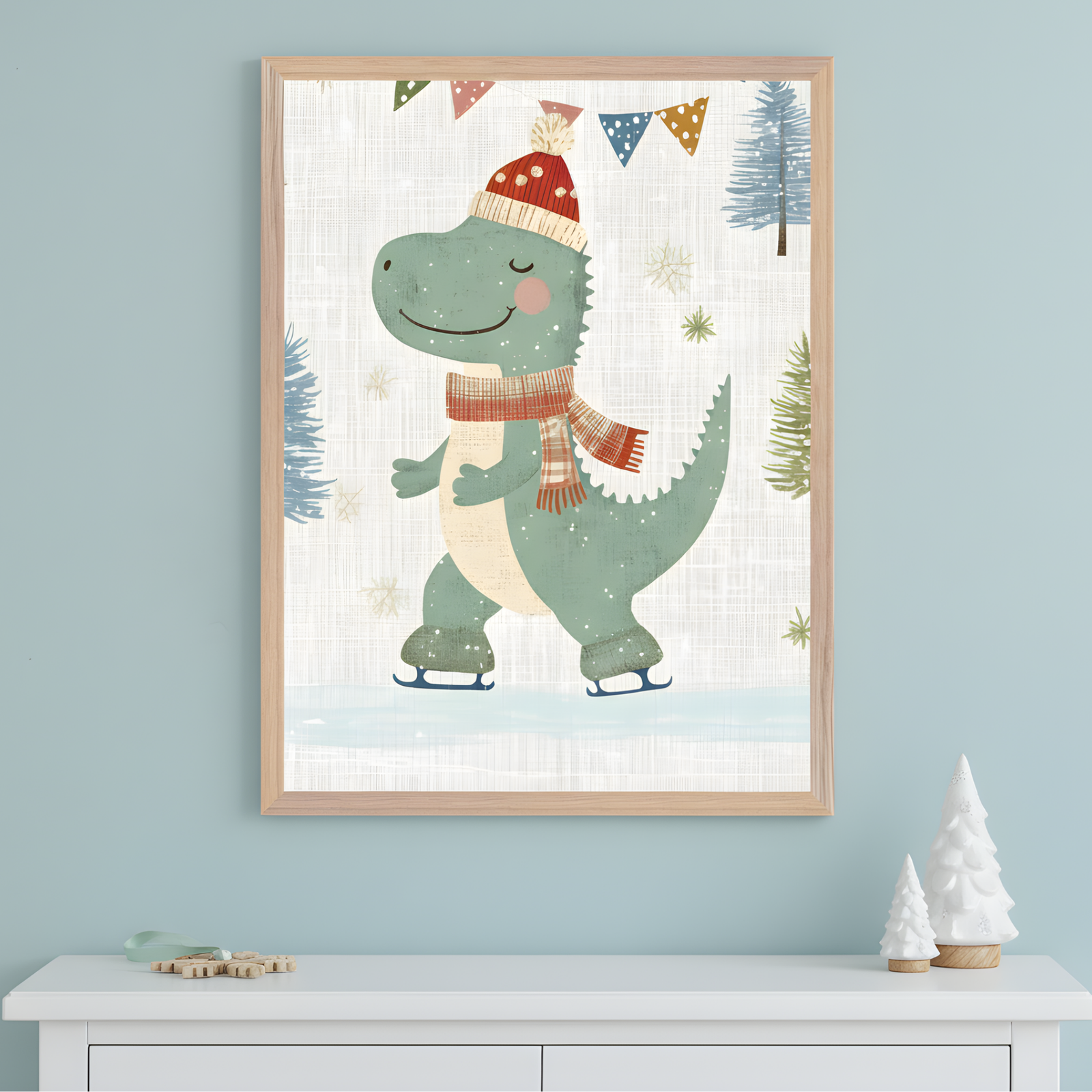 A framed winter-themed illustration shows a cheerful green dinosaur ice skating, wearing a red beanie with a pom-pom and a striped scarf. The background features snowflakes, evergreen trees, and festive bunting, with white ceramic Christmas trees and wooden snowflake ornaments on the dresser below.