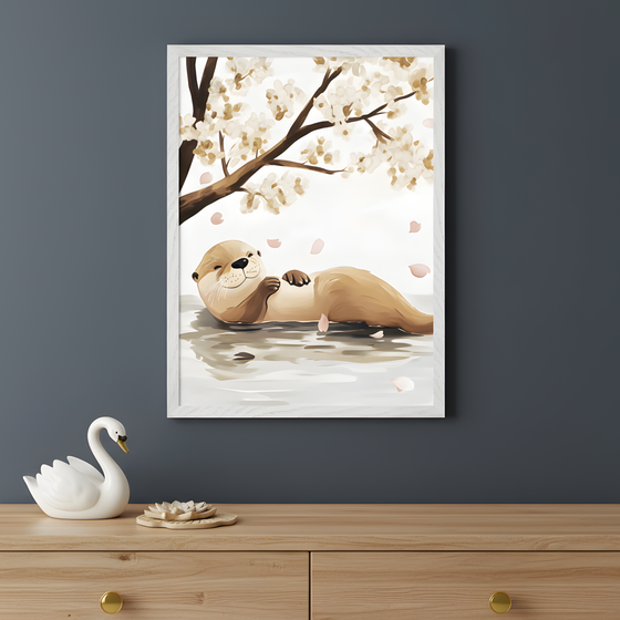 Framed wall art of a relaxed otter floating on its back in calm water, gazing up with a gentle smile as pink flower petals drift down from a blossoming tree above. The background features soft white and beige tones, creating a peaceful, springtime feel. A ceramic swan and a lotus-shaped dish sit on the wooden dresser beneath, enhancing the tranquil scene.