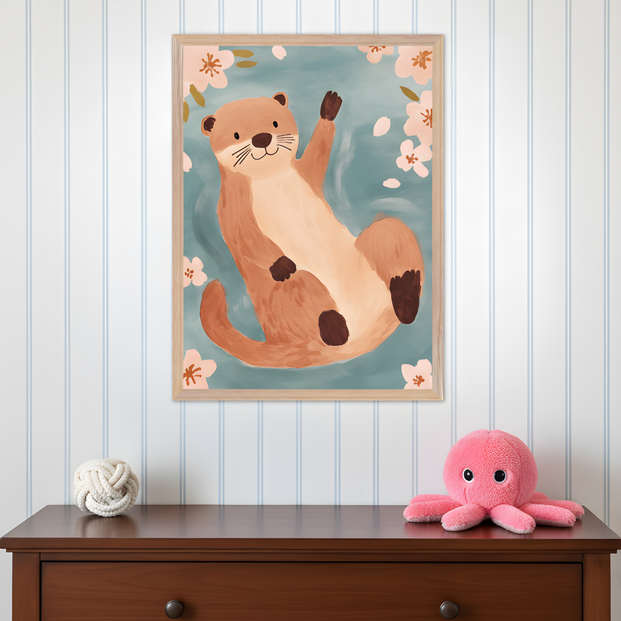 A framed illustration of a cheerful brown otter floating on its back in blue water, surrounded by pink blossoms and leaves. The playful scene hangs above a wooden dresser topped with a white rope knot decor and a bright pink octopus plush toy.