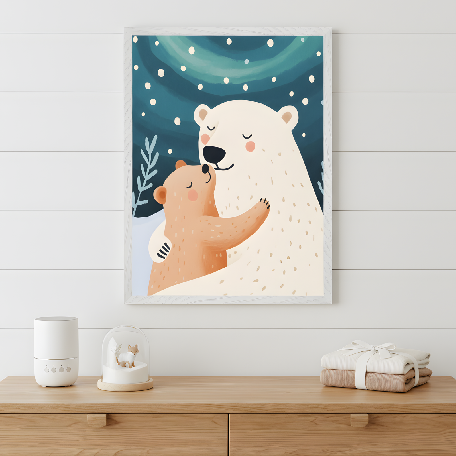 Framed nursery art of a warm embrace between a polar bear and a brown bear cub, set against a snowy night sky with falling snowflakes and swirling auroras. The bears have peaceful expressions, creating a cozy, loving vibe. The light wood dresser below is styled with folded baby clothes, a white diffuser, and a snow globe featuring a tiny fox.