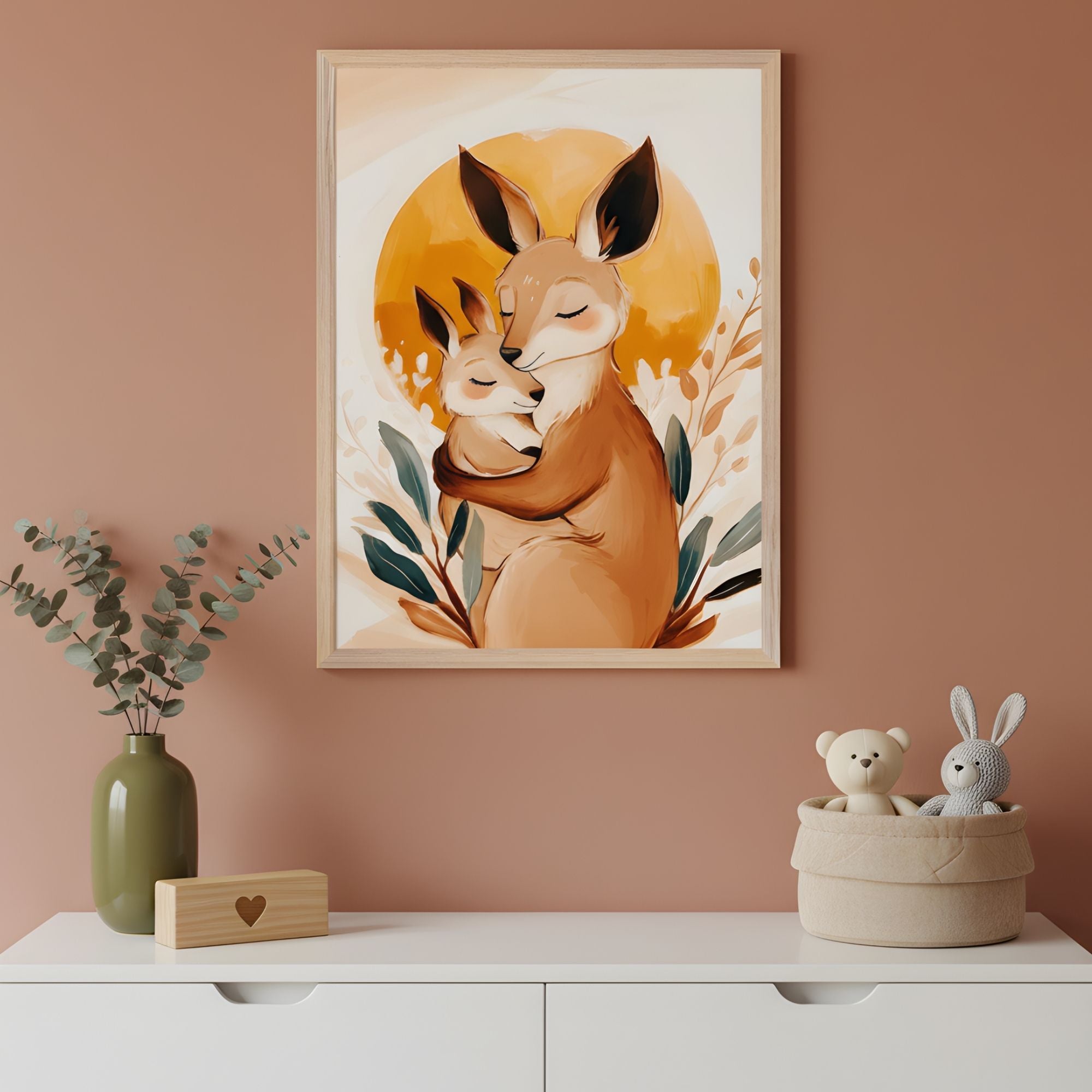 A framed illustration features a tender scene of a kangaroo hugging its joey, both with eyes closed, set against a warm golden sun and surrounded by leafy plants. The artwork hangs above a white dresser with a green vase, a wooden block with a heart cutout, and a soft basket holding plush bear and bunny toys.