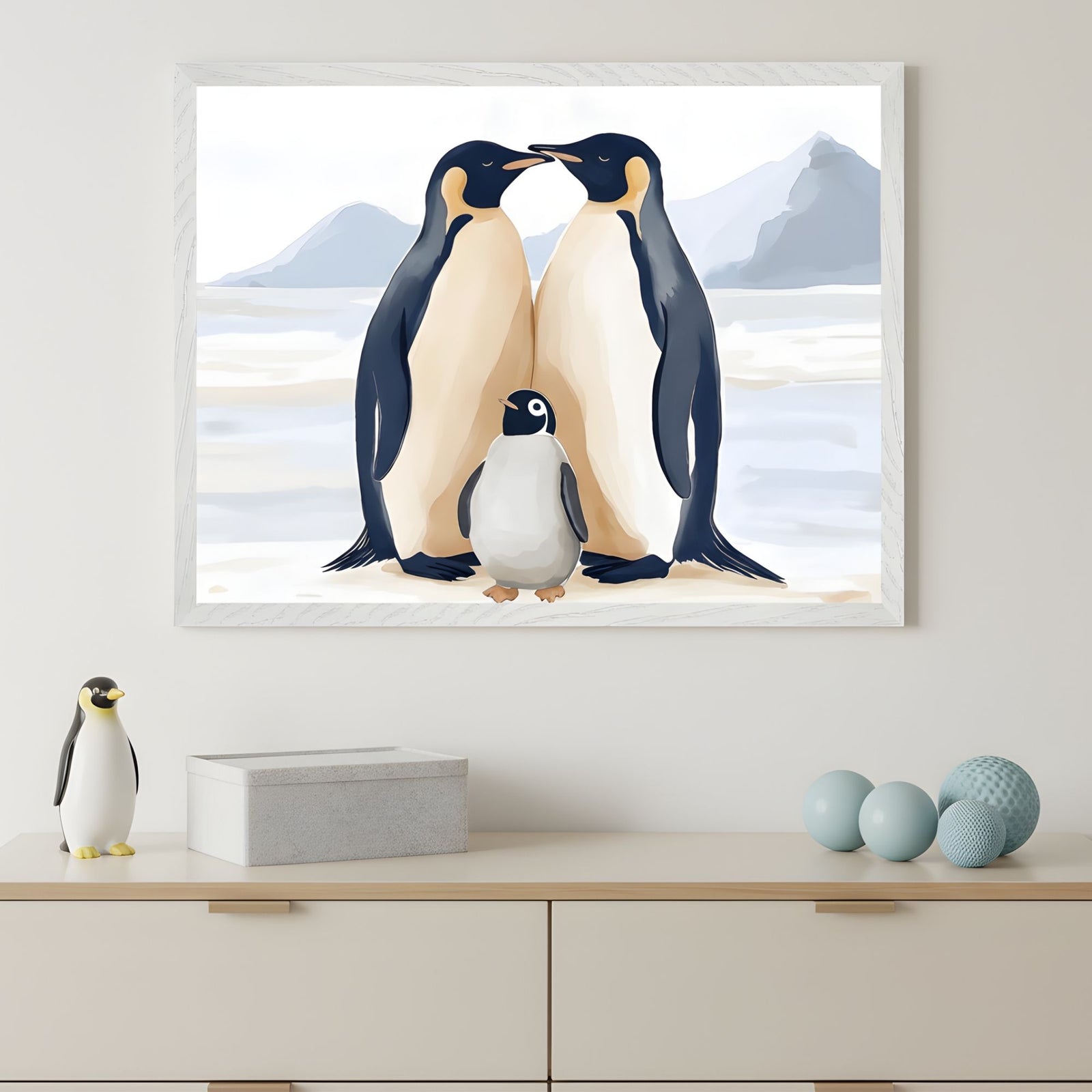 Framed illustration of two adult emperor penguins standing close together with a fluffy penguin chick between them, set against a snowy Antarctic backdrop with distant mountains. The print hangs above a minimalist dresser decorated with a penguin figurine, a concrete box, and blue decorative spheres, creating a clean and modern nursery vibe.