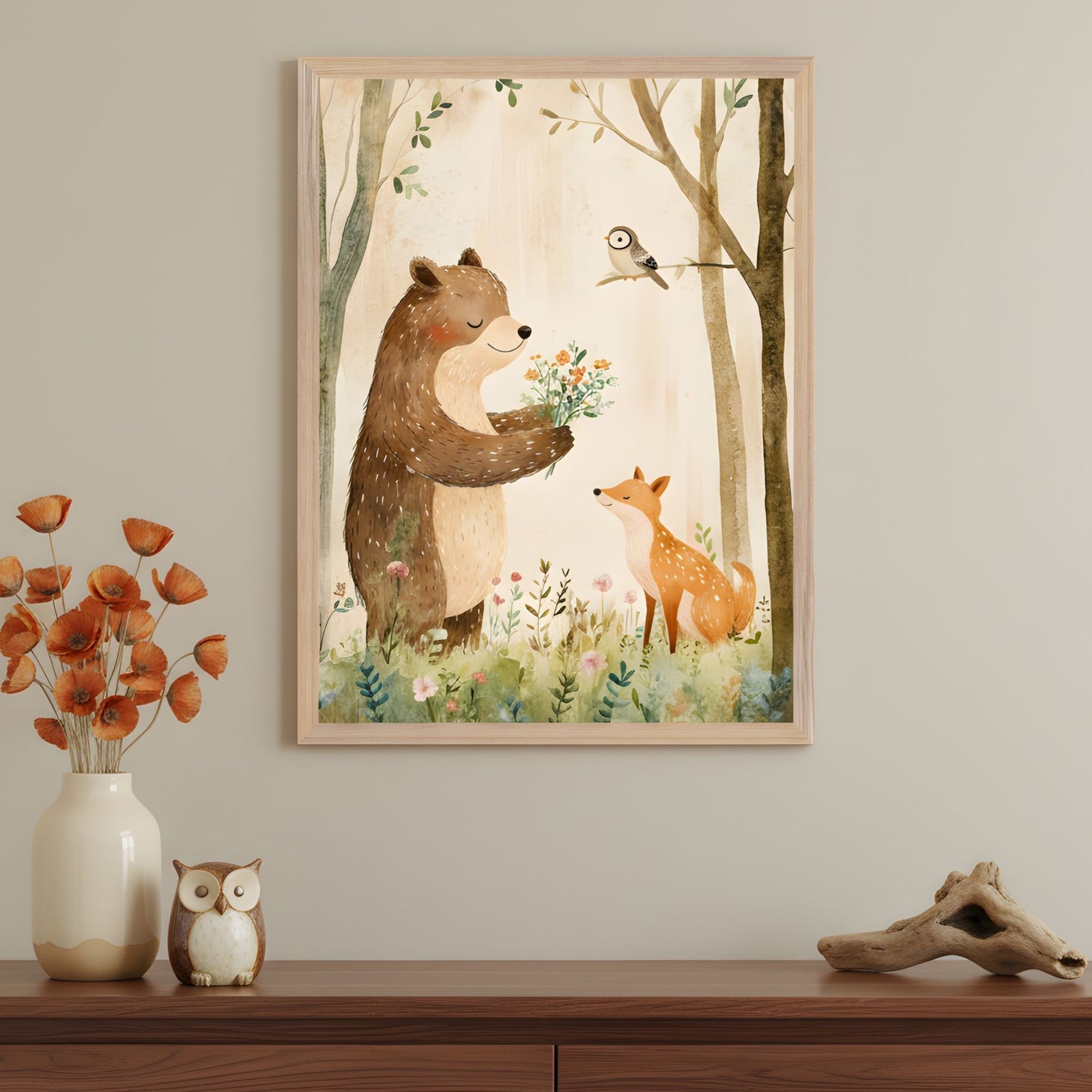 A framed woodland-themed illustration shows a friendly bear offering a bouquet of wildflowers to a fox, with a bird perched on a tree branch above them. The artwork hangs on a beige wall above a wooden sideboard decorated with a ceramic owl, a vase of orange poppies, and a piece of driftwood.