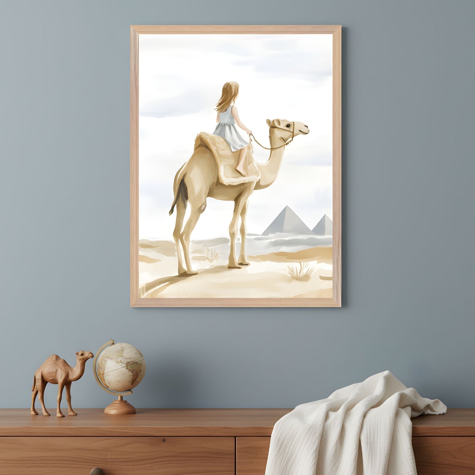A framed illustration of a young girl in a light blue dress riding a camel in a desert landscape, looking toward the pyramids in the distance. The artwork hangs above a wooden dresser decorated with a small globe, a carved wooden camel, and a draped white cloth.