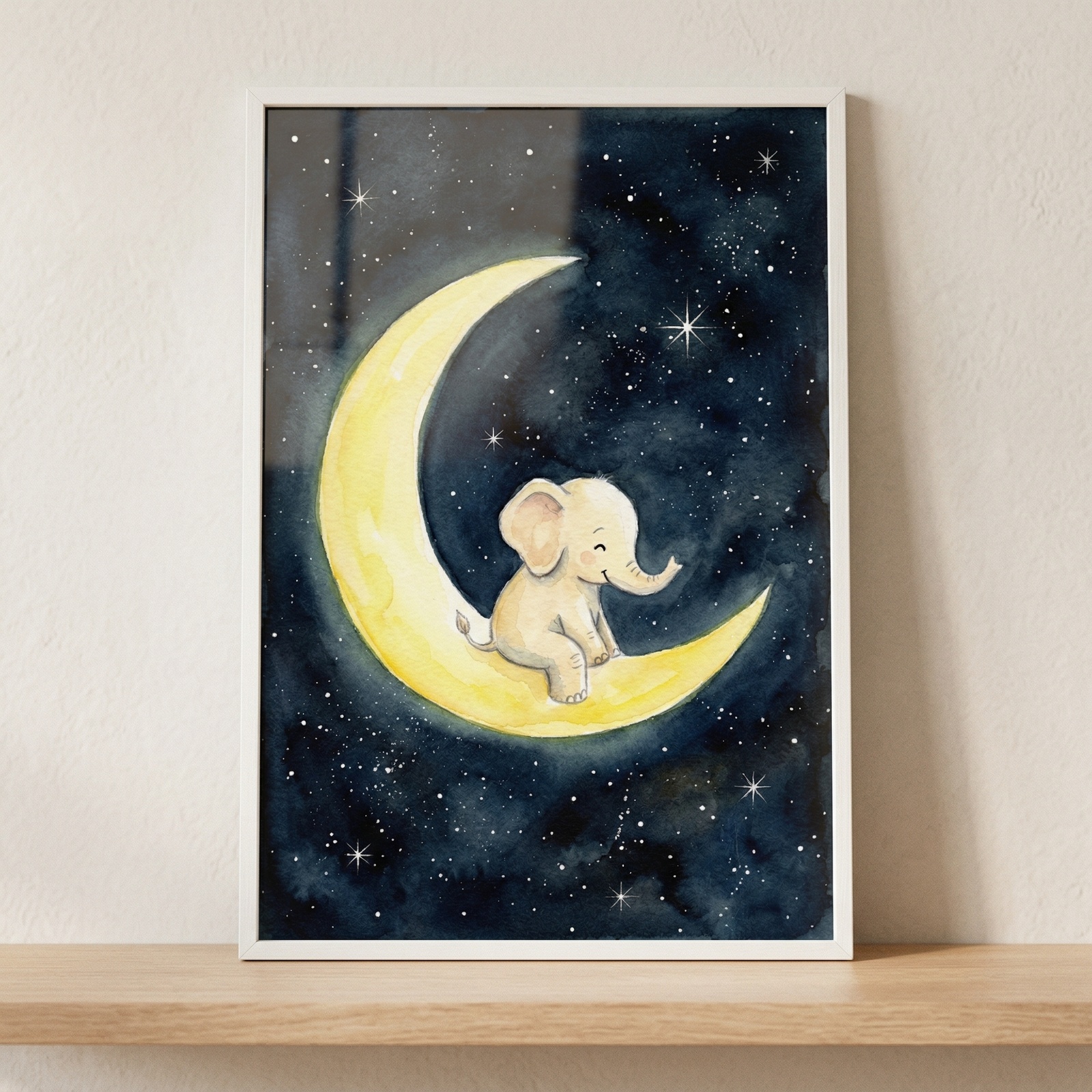 A watercolor-style illustration of a small, smiling baby elephant sitting on a bright yellow crescent moon. The moon is set against a dark blue night sky filled with many small, twinkling white stars of varying sizes. The artwork is enclosed in a simple white rectangular frame and is displayed resting on a light-colored wooden shelf against a neutral, textured wall.