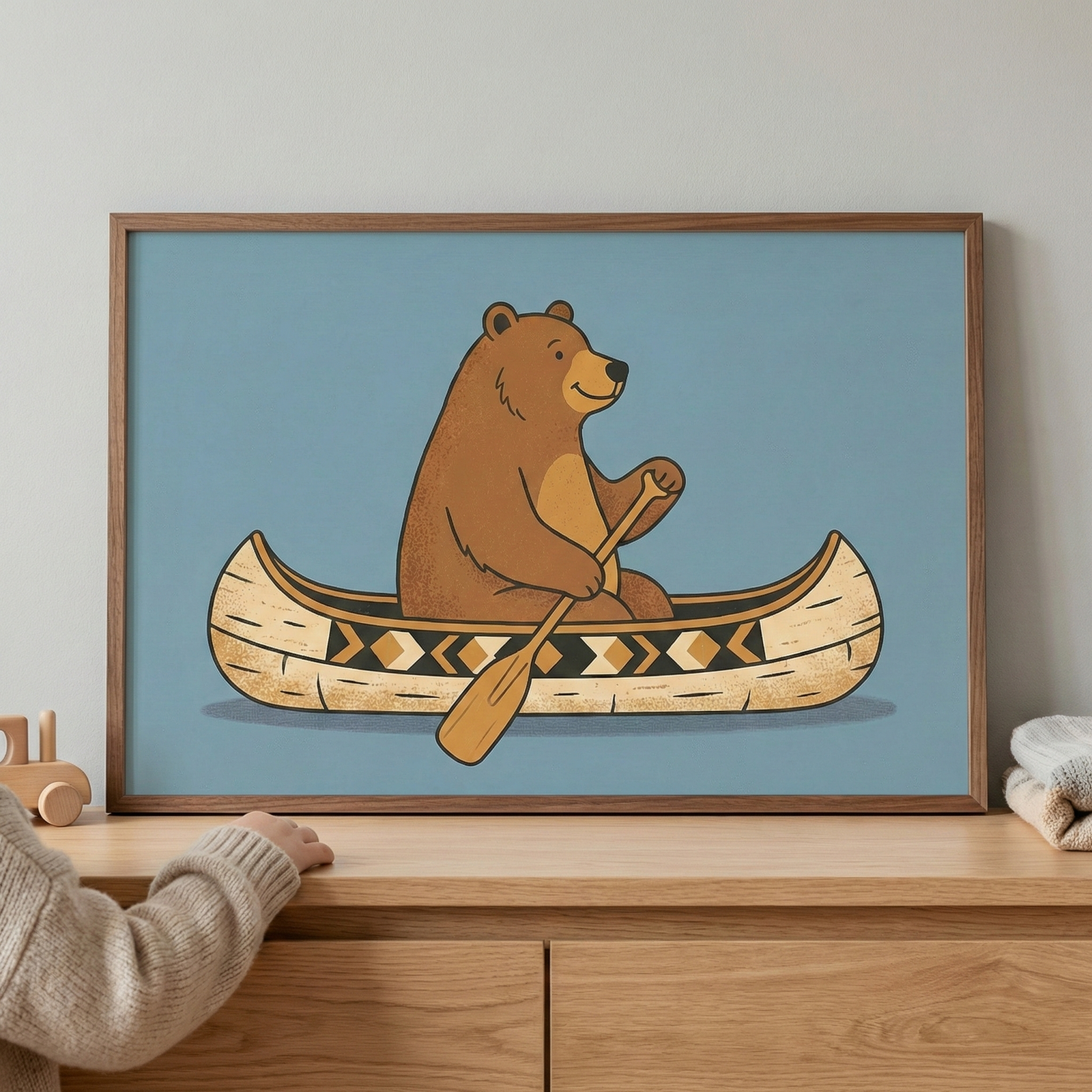 Framed woodland animal wall art featuring a bear in a canoe, displayed in a brown wooden frame on a light wood dresser next to a child's hand and a wooden toy.
