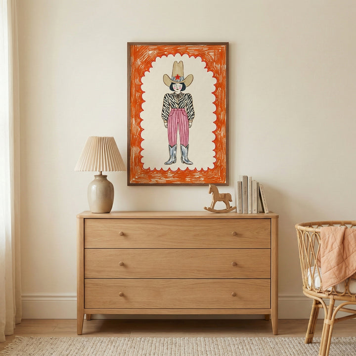 Warm and neutral-toned nursery featuring a framed cowboy-themed artwork above a wooden dresser. The colorful illustration shows a stylized cowboy with a zebra-print top and pink pants, bringing a fun and modern western vibe to the space.