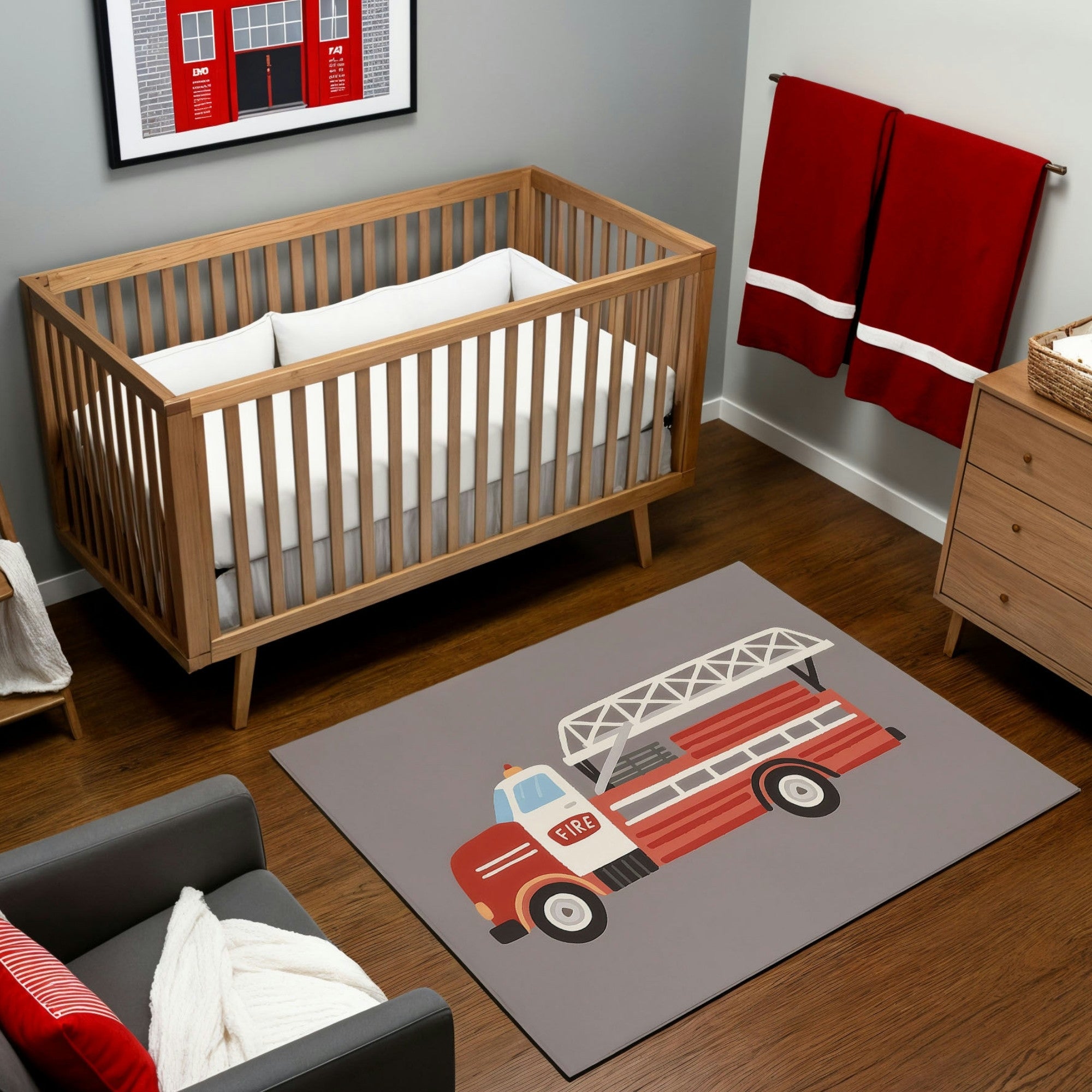 A firetruck-themed nursery featuring a natural wood crib and dresser on dark wood flooring, accented by red towels and a framed firehouse print on the gray wall. The room includes a whimsical area rug with a bright red firetruck illustration, complete with a ladder and “FIRE” text. A cozy gray armchair with a red striped pillow and knit throw enhances the modern and playful firehouse-inspired design.