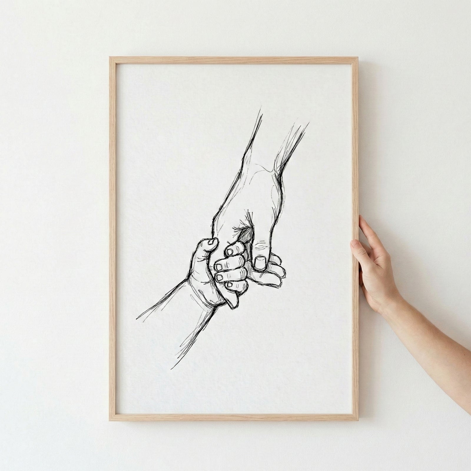 Framed wall art featuring a pencil-style sketch of a parent and child holding hands, held by a person's hand against a white wall.