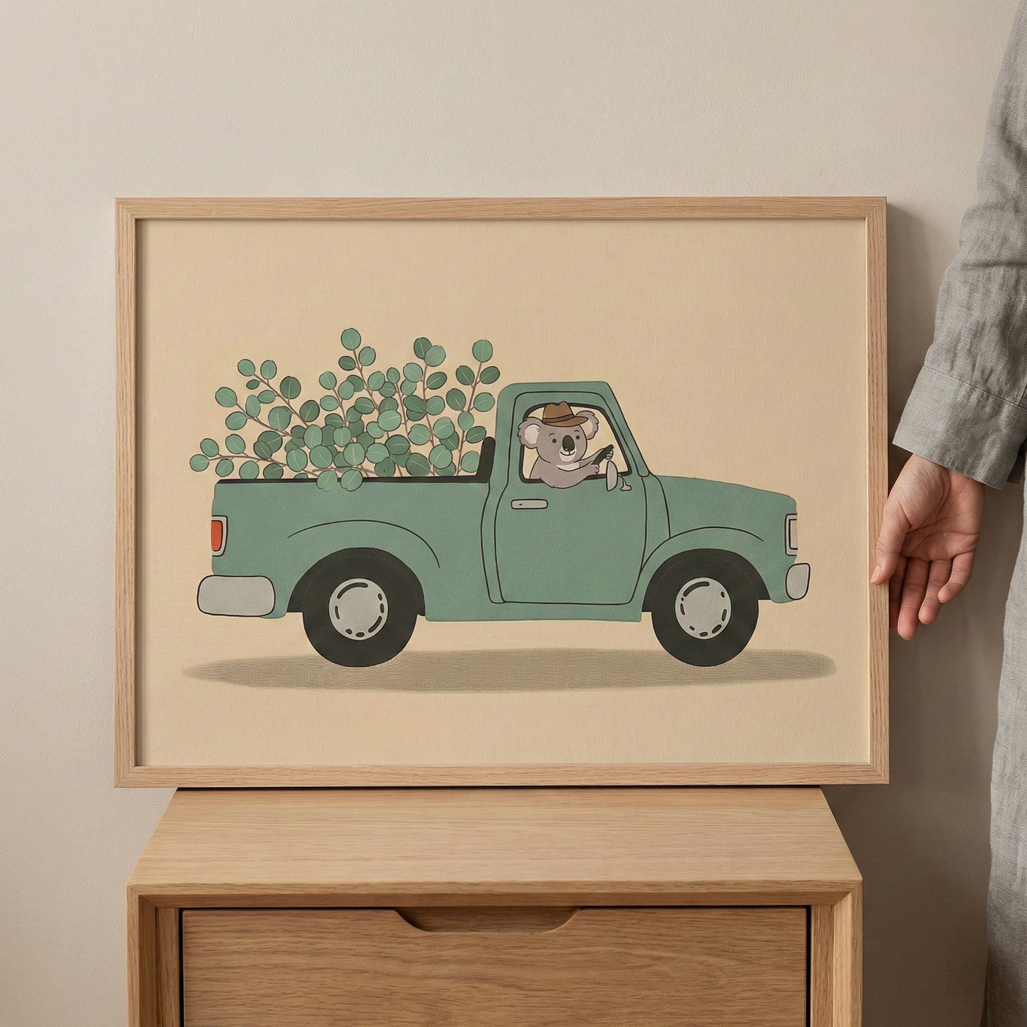 Framed koala driving a green car filled with eucalyptus wall art in a light wood frame, held by a hand against a neutral wall and resting on a wooden cabinet.