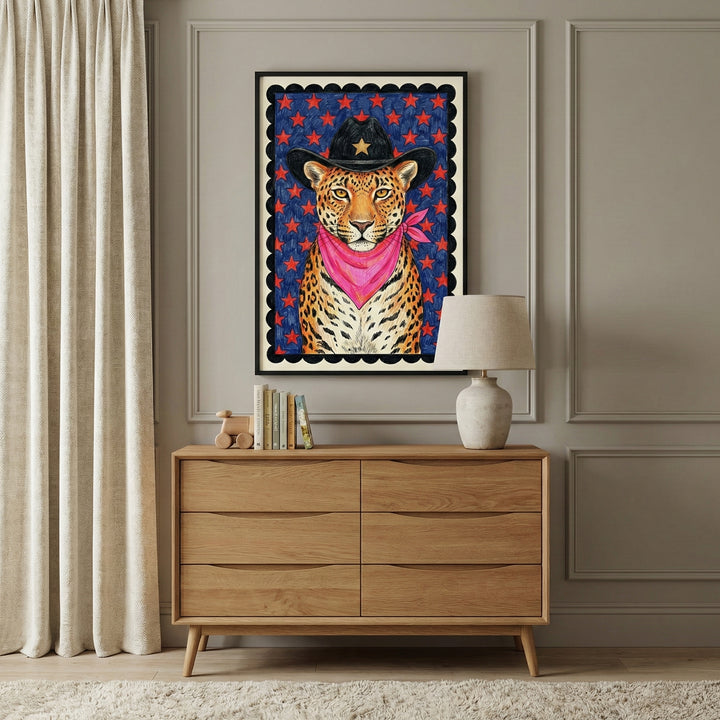 A framed illustration of a leopard wearing a black cowboy hat with a gold star and a bright pink bandana hangs above a modern wooden dresser. The room features elegant wall molding, neutral curtains, and a soft white rug.