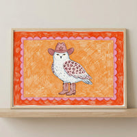 Framed cowboy owl animal wall art in a light wood frame sitting on a wooden shelf against a neutral wall.