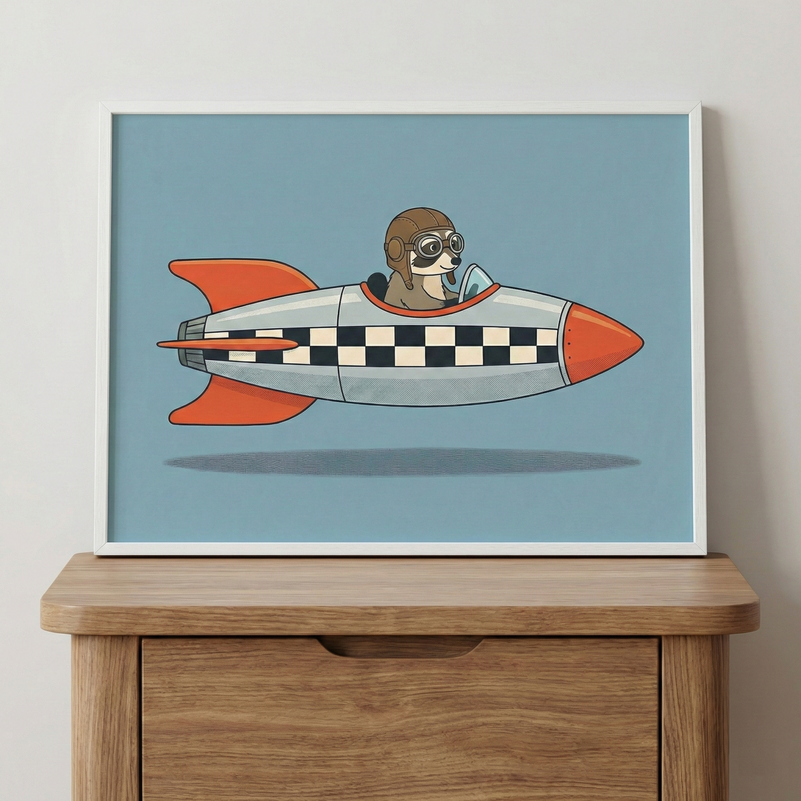 Framed raccoon in a space rocket wall art sitting in a thin white frame atop a light-toned wooden cabinet against a plain white wall.