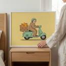 Framed sloth wall art featuring a sloth on a green scooter with a crate of mango fruit, held by a hand in a white frame resting on a wooden nightstand.