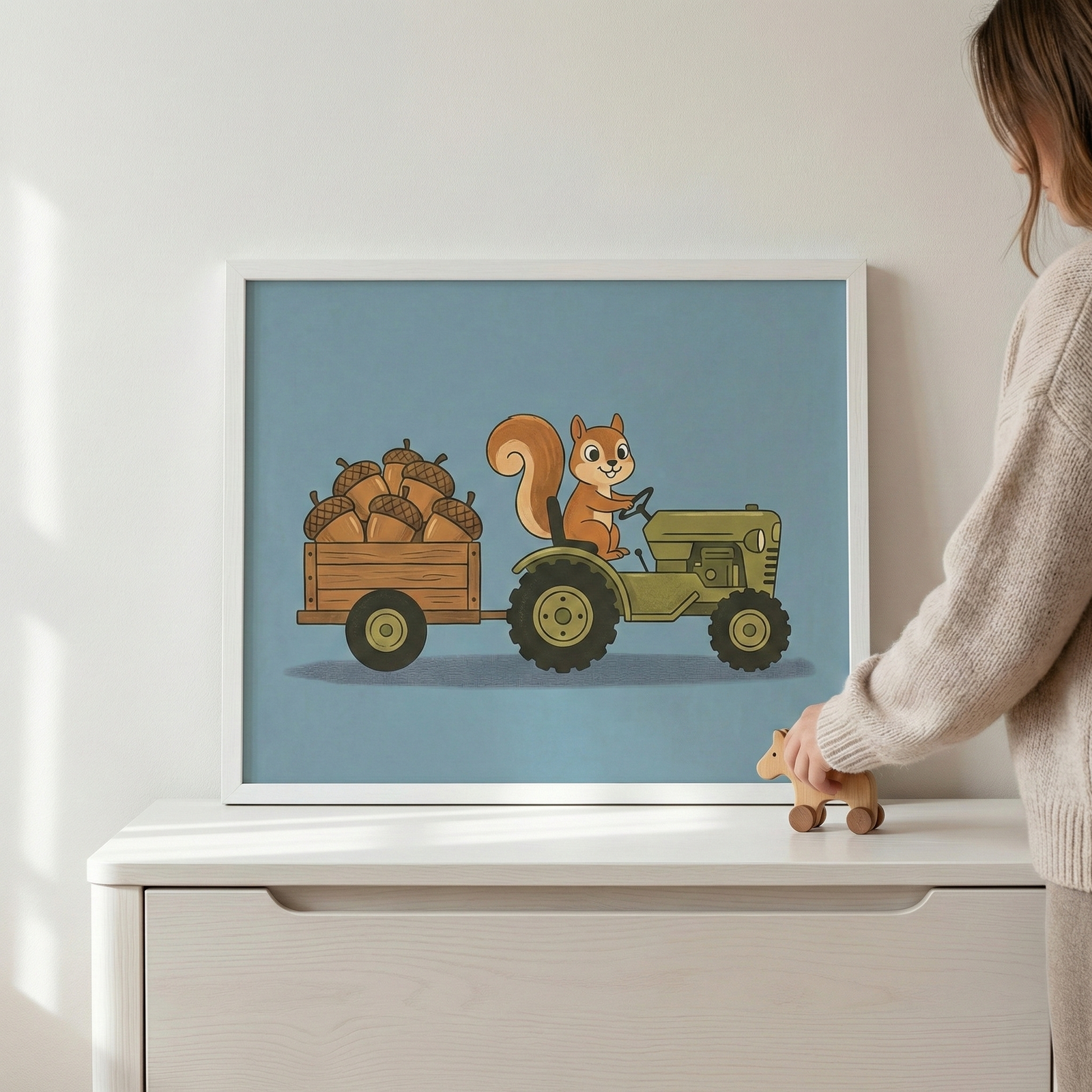 Framed wall art featuring a squirrel driving a green tractor pulling a trailer of acorns, presented in a white frame on a white dresser with a person's hand touching a wooden toy.