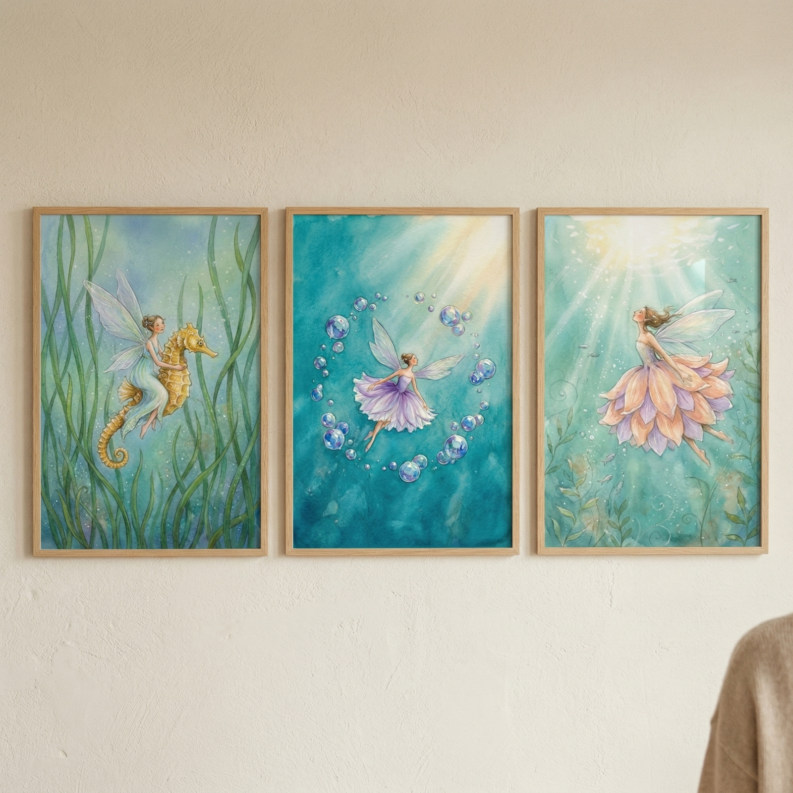 Three framed watercolor-style illustrations hang on a beige wall, featuring magical underwater scenes. The left artwork shows a fairy with pale blue wings riding a golden seahorse through tall green seaweed. The center piece depicts a fairy in a purple dress surrounded by a ring of bubbles in deep blue water. The right illustration features a fairy with large petal-like wings in shades of pink and orange, floating towards light filtering from the surface of clear teal water.