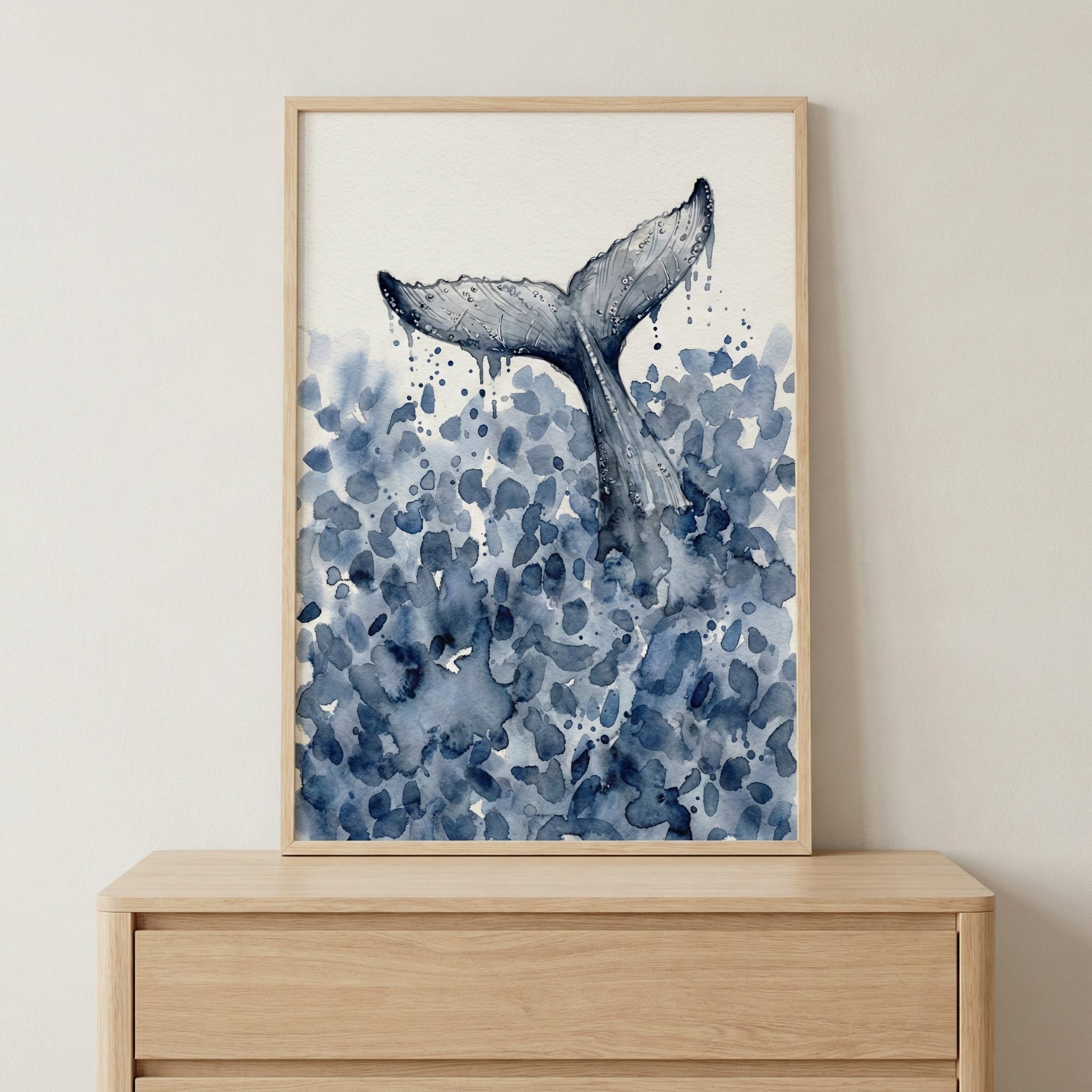 Close-up of a watercolor painting showing a whale’s tail splashing into the sea, with deep blue brushstrokes mimicking coastal waters. The art is framed in light wood and styled simply atop a natural-toned dresser.