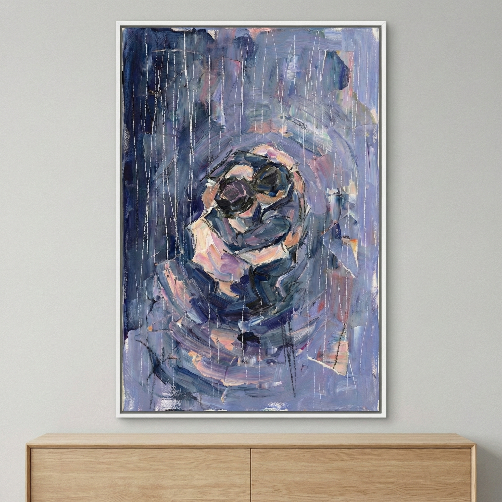 This vertical artwork captures two abstract figures in a tight embrace at its center. Rendered with expressive, thick strokes of navy, lavender, and pale pink, the forms emerge from a background of layered blues and purples. Delicate, vertical white lines are drawn across the surface, evoking a sense of falling rain. The textured piece is presented in a slim white frame, hanging on a neutral wall above a light-toned wooden chest of drawers. The style is emotive and gestural.