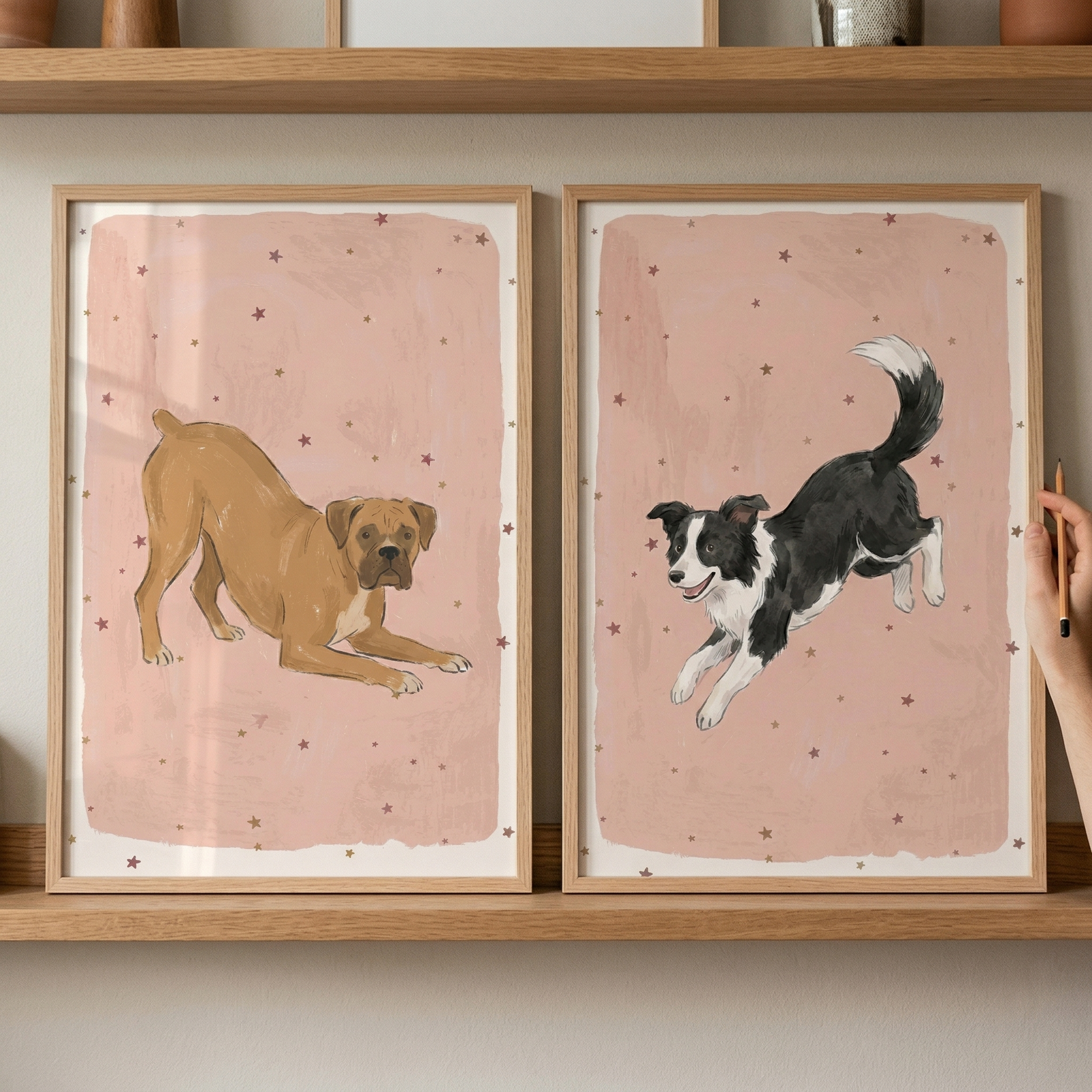 Two framed dog illustrations sit side-by-side on a light wood shelf. Both have a muted pink background decorated with small, dark red stars. The illustration on the left shows a short-haired, tan-colored dog in a playful crouching pose. The illustration on the right depicts a black and white dog with a fluffy tail mid-leap. 