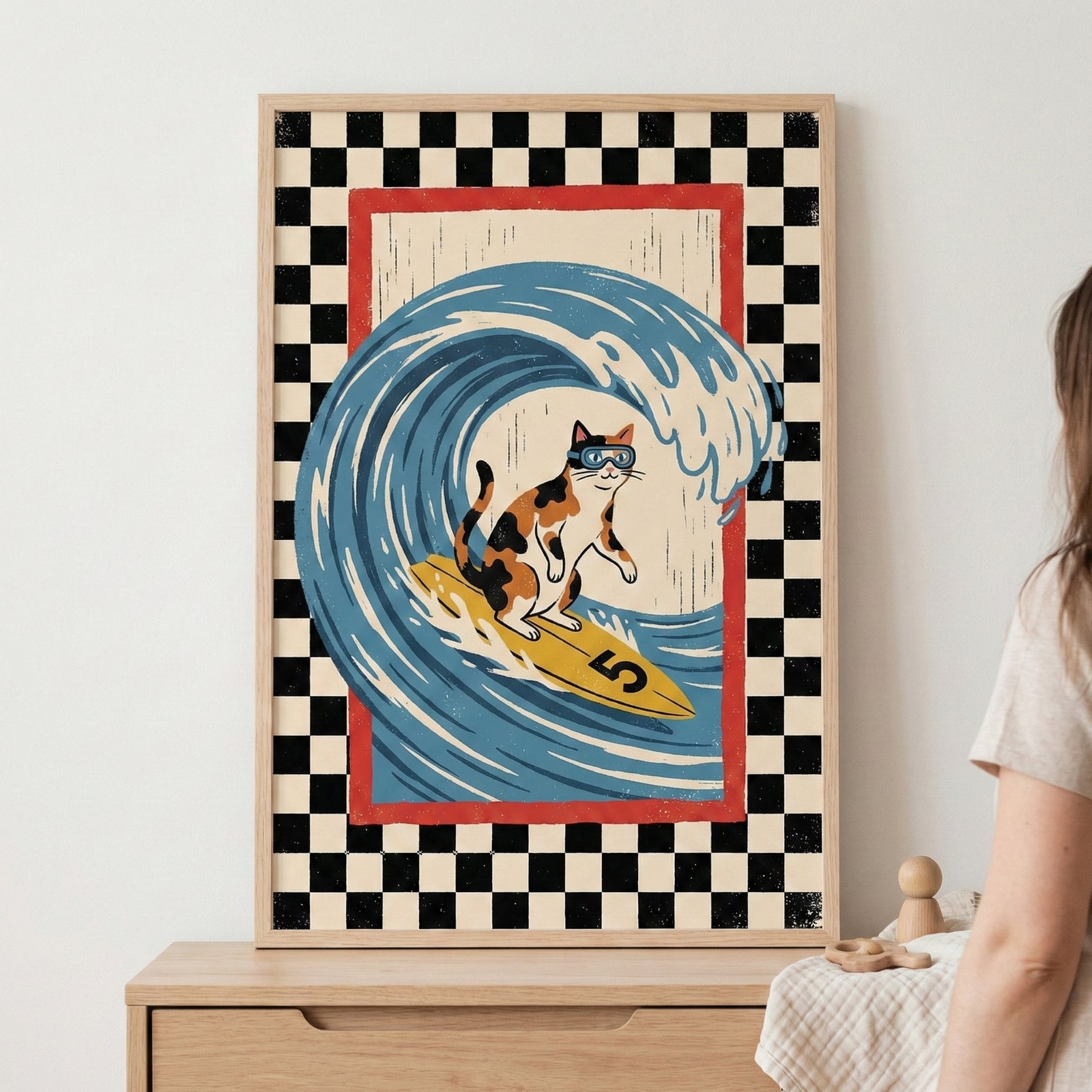 A framed graphic illustration of a calico cat wearing blue swimming goggles and surfing on a yellow surfboard with the number "5". The cat is positioned inside the curve of a large, stylized blue and white ocean wave. The central scene is enclosed by a thin red border, which is itself surrounded by a wide black and white checkerboard pattern. The framed artwork is propped up on a light-colored wooden dresser, with the shoulder of a person and some small wooden objects visible in the foreground.