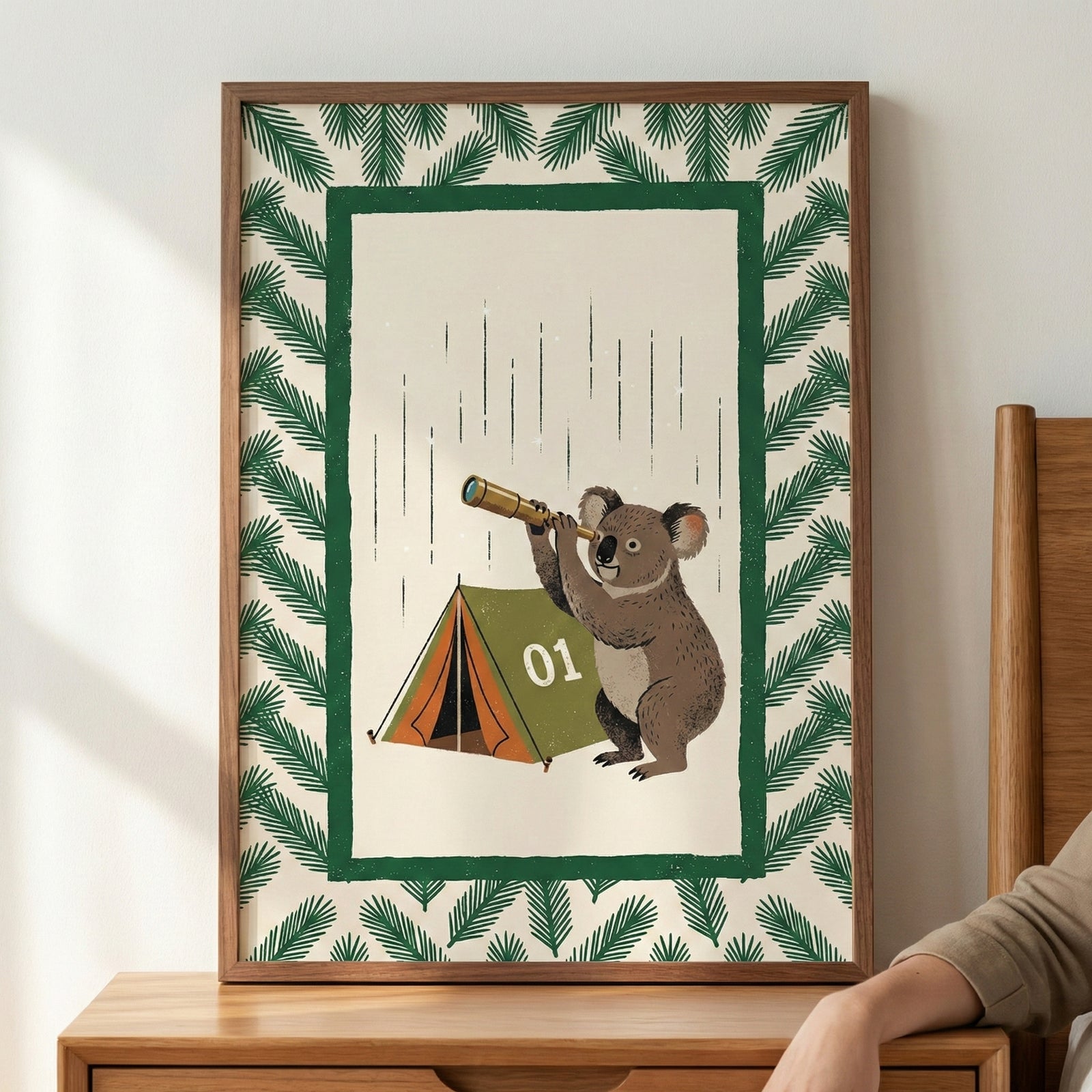 An illustration in a wooden frame sits on a wooden surface against a plain wall. The artwork features a koala standing next to a small green and orange camping tent marked with the number "01." The koala is holding a small handheld telescope and looking through it. The central scene is set against a light beige background with thin, vertical dashed lines and is framed by a solid green rectangular border. Surrounding this inner border is a pattern of stylized green pine-like branches.