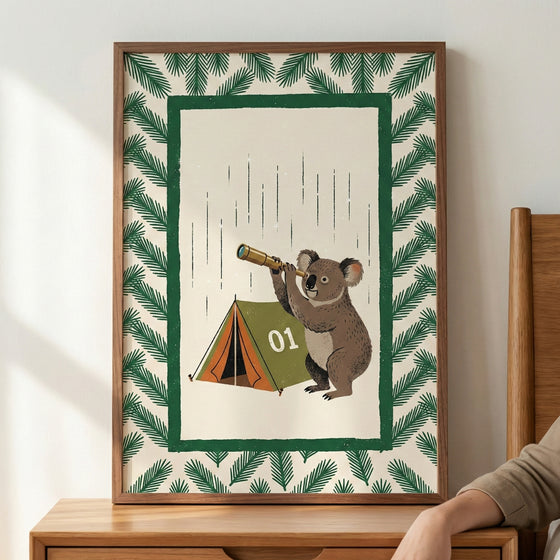 An illustration in a wooden frame sits on a wooden surface against a plain wall. The artwork features a koala standing next to a small green and orange camping tent marked with the number "01." The koala is holding a small handheld telescope and looking through it. The central scene is set against a light beige background with thin, vertical dashed lines and is framed by a solid green rectangular border. Surrounding this inner border is a pattern of stylized green pine-like branches.
