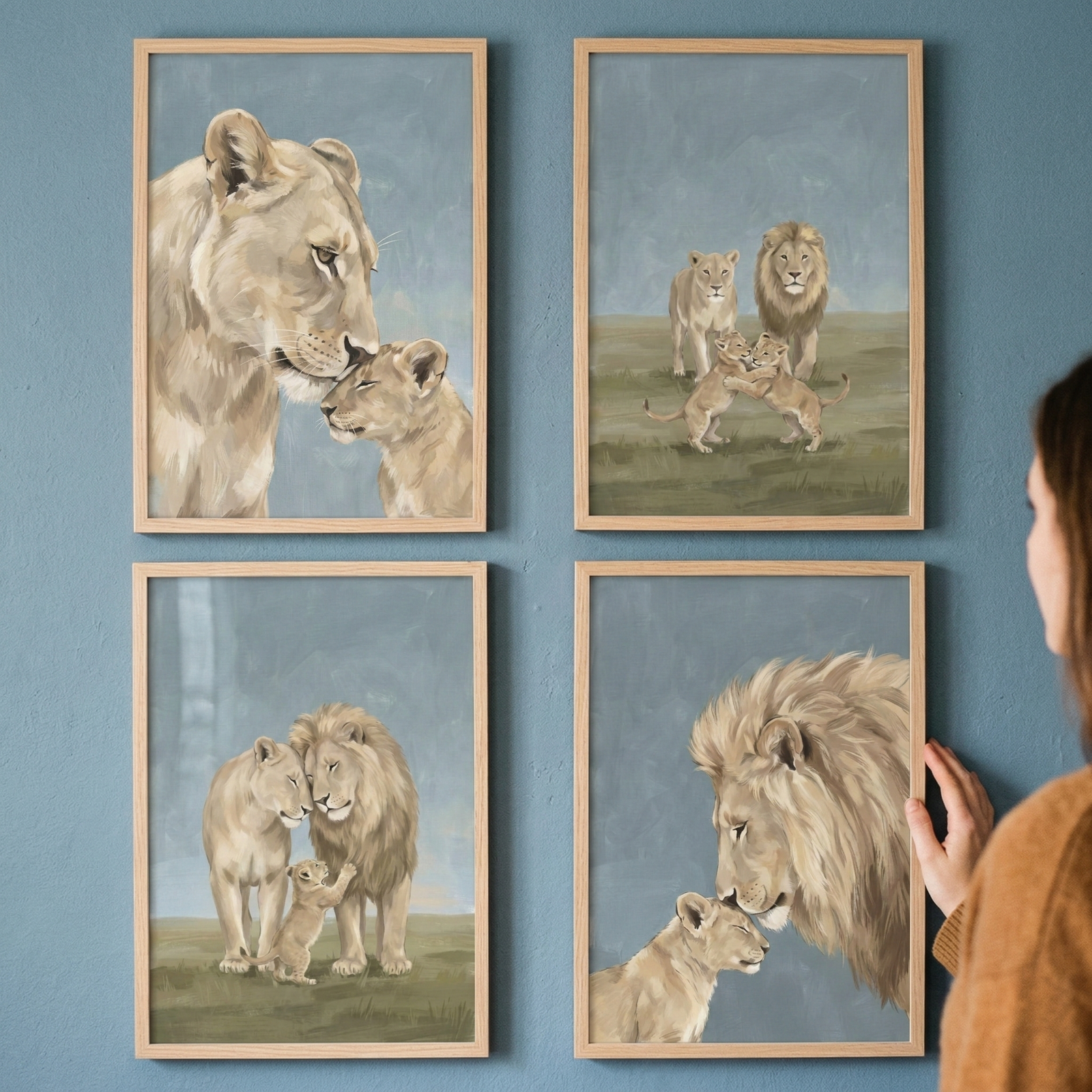 A person hangs four framed illustrations on a blue wall, each depicting a different lion family scene. The artwork uses soft, muted tones of beige and blue with a gentle, blended style. One frame shows a lioness nuzzling her cub, while another shows a male lion with a full mane interacting with a young one. Other scenes include full families of lions together in grassy landscapes. The pieces are encased in light-colored wooden frames. A woman’s hand is visible as she adjusts the bottom-right frame.