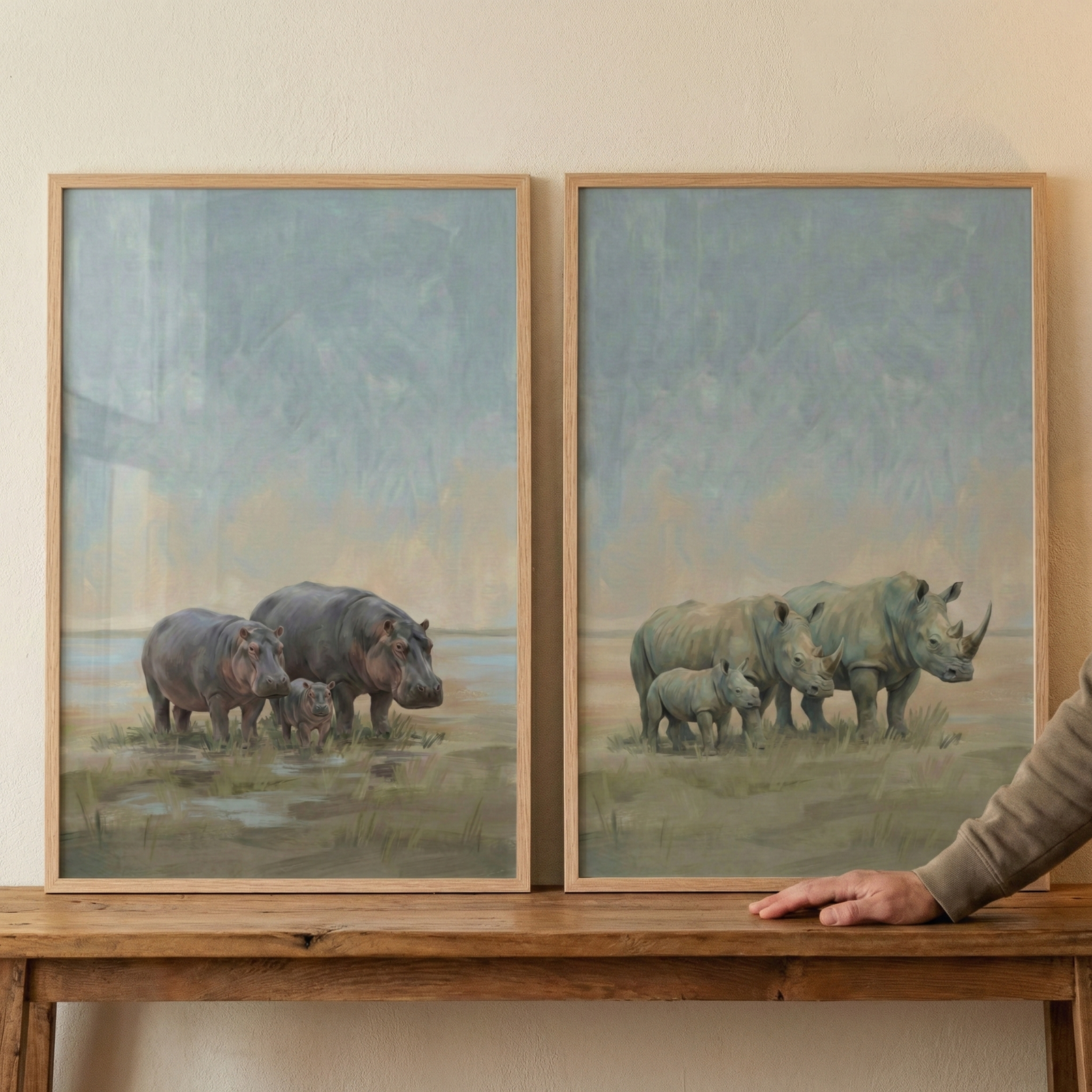 Two rectangular art pieces in light wood frames sit side-by-side on a rustic wooden table. The left illustration features three hippopotamuses—two adults and a calf—in a shallow wetland. The right illustration depicts three rhinoceroses—two adults and a calf—on a grassy field. Both pieces have a soft, impressionistic style with a muted palette of grays, blues, and earth tones. A hand rests on the table's edge beside the frames.