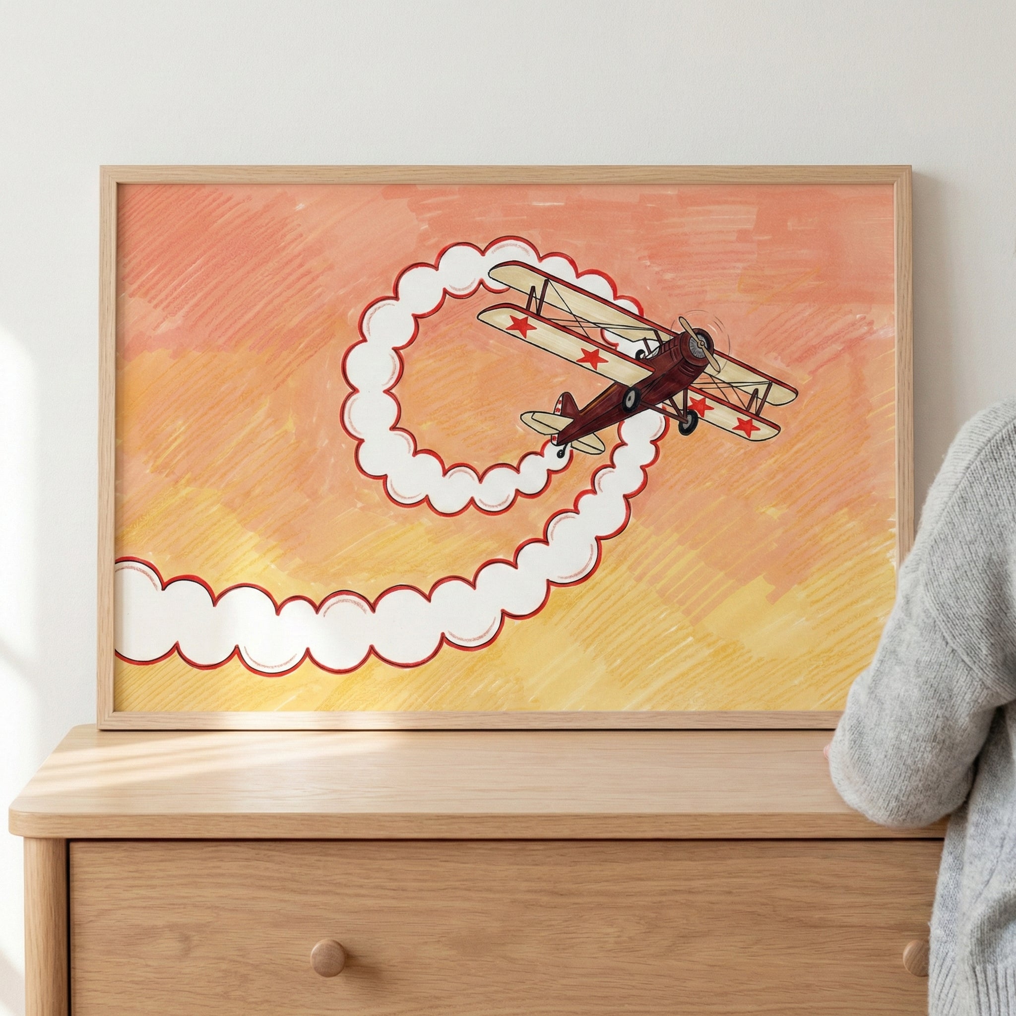Framed artwork of a vintage red and cream biplane with red star markings, creating a loop of fluffy white smoke against a warm orange and yellow sunset background, resting on a wooden dresser.