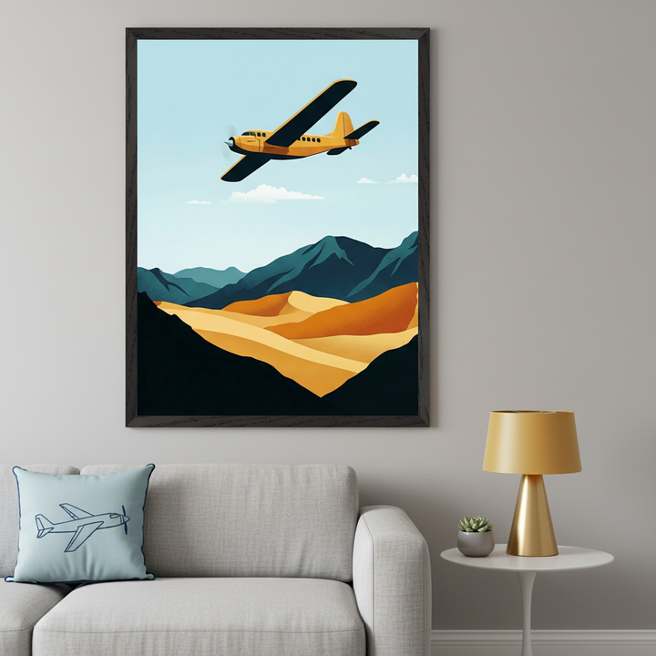 Modern living room with a large framed artwork hanging above a gray sofa. The art features a minimalist illustration of a yellow biplane flying over orange sand dunes and blue-green mountains under a light blue sky. A light blue pillow with a plane outline rests on the sofa, and a gold lamp is on a white side table.