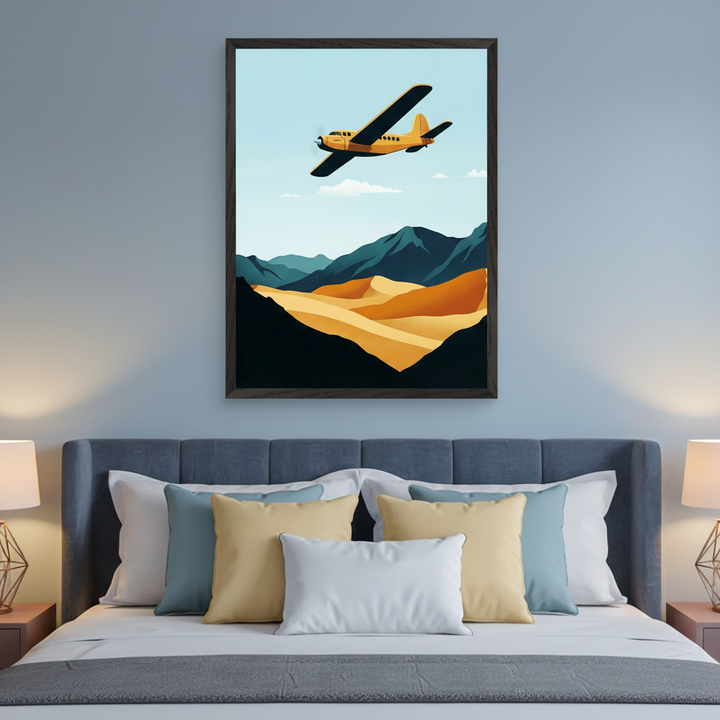 Modern bedroom with a grey upholstered bed. A large framed print featuring a yellow plane flying over teal mountains and golden sand dunes in a minimalist style hangs centered above the bed.