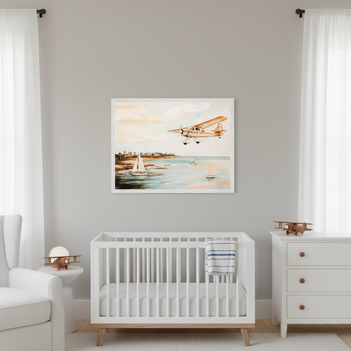 Modern nursery with white crib, dresser, and armchair. A large framed watercolor-style print of a golden airplane flying over a calm ocean with sailboats and a tropical coastline hangs above the crib.