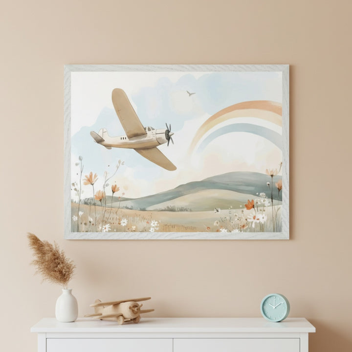Nursery with a large framed watercolor print hanging above a white dresser. The print features a beige airplane flying over a floral field and hills with a rainbow in the sky. A wooden airplane toy, vase, and clock sit on the dresser.