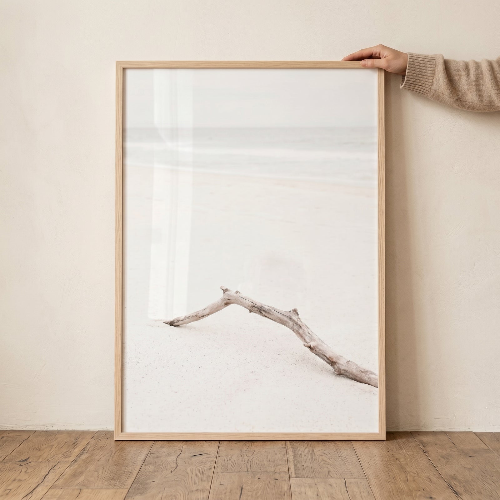 Airy white sand driftwood wall art in a light wood frame being held against a neutral wall on a wooden floor.