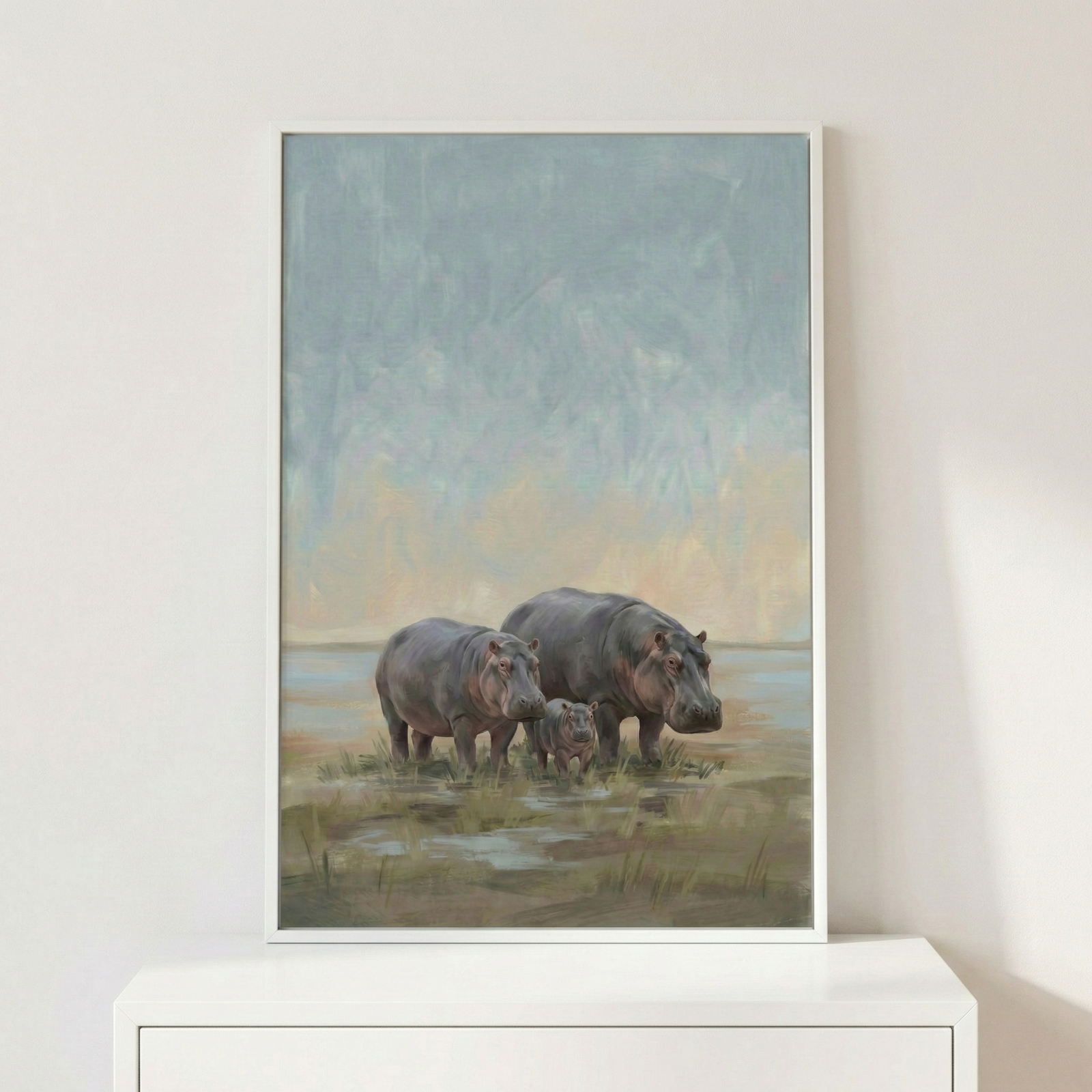 A vertically oriented artwork in a thin white frame depicts a family of three hippopotamuses standing in a marshy landscape. Two larger hippopotamuses flank a smaller calf in the center. The scene is set against an atmospheric background of a textured, soft-toned sky in shades of blue, grey, and pale yellow. The hippos stand among low grasses and shallow pools of water. The framed piece is displayed on a white flat surface against a plain white wall.