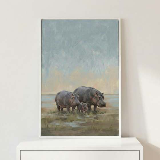 A vertically oriented artwork in a thin white frame depicts a family of three hippopotamuses standing in a marshy landscape. Two larger hippopotamuses flank a smaller calf in the center. The scene is set against an atmospheric background of a textured, soft-toned sky in shades of blue, grey, and pale yellow. The hippos stand among low grasses and shallow pools of water. The framed piece is displayed on a white flat surface against a plain white wall.