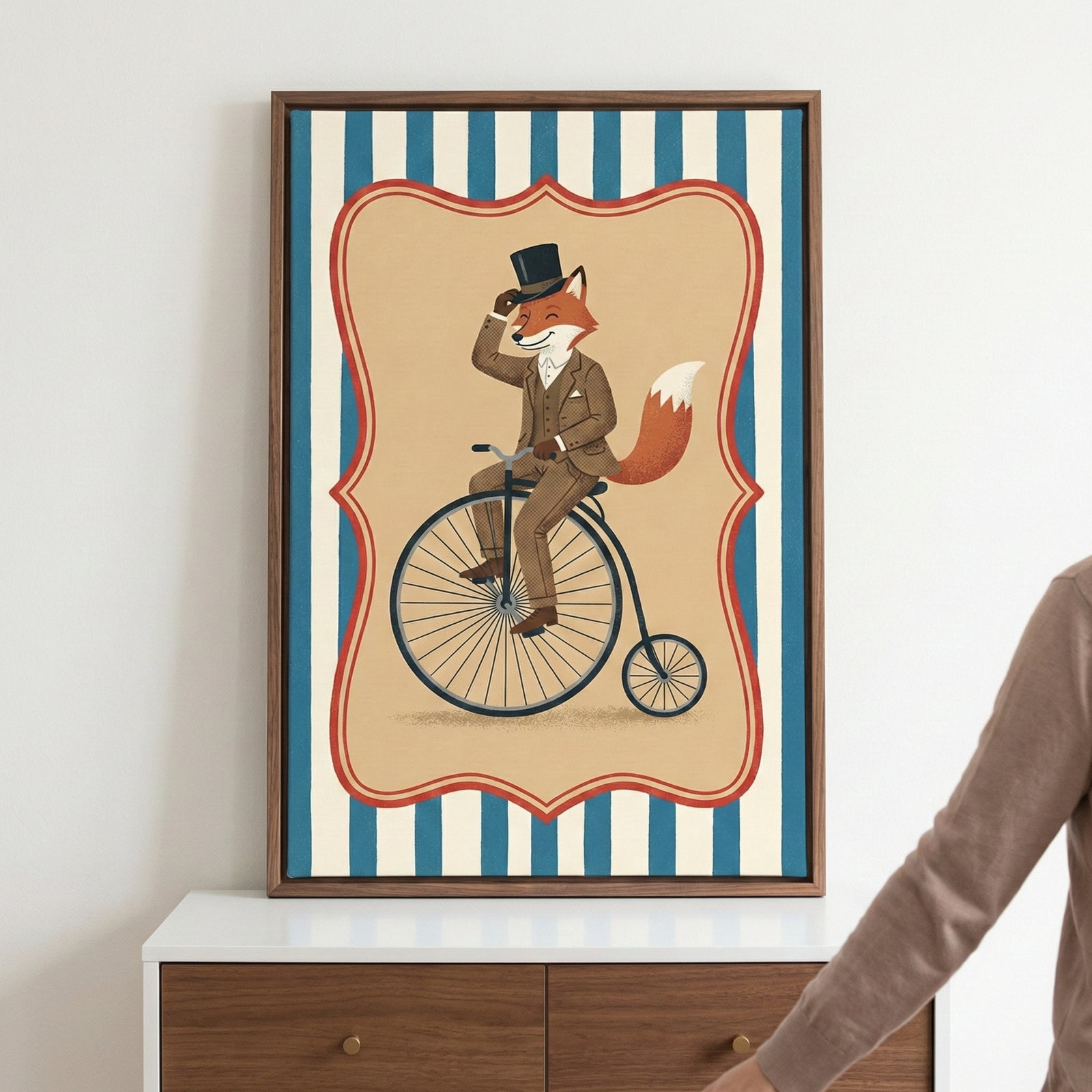 A framed vertical illustration of an anthropomorphic fox dressed in a brown three-piece suit and a black top hat. The fox is depicted riding a black penny-farthing bicycle. This central scene is enclosed in a tan, ornate-bordered frame, set against a background of bold blue and white vertical stripes. The artwork is displayed in a dark wood frame, resting on a white and brown wooden dresser against a plain white wall. A person's arm is partially visible on the right side of the image.