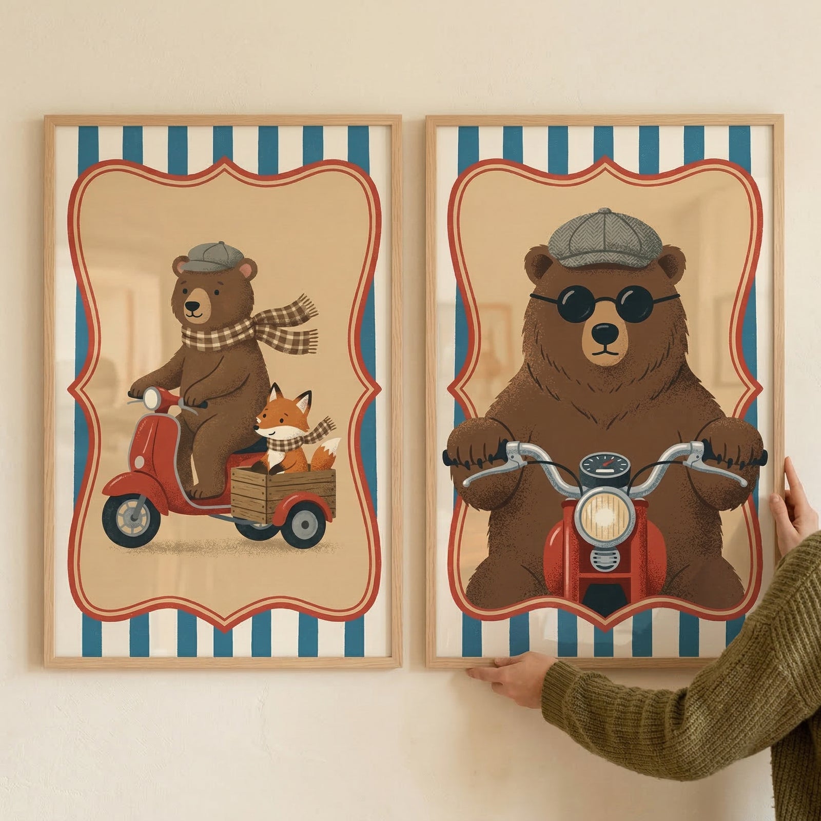 Two framed illustrations hang on a white wall. The left image shows a brown bear in a grey flat cap and checkered scarf riding a red scooter with a small fox in a rear wooden crate. The right image depicts a large brown bear wearing round sunglasses and a flat cap, riding a red motorcycle head-on. Both artworks feature a scalloped border against blue and white stripes. A person’s hands, wearing a green sweater, are seen holding the bottom edge of the frame on the right.