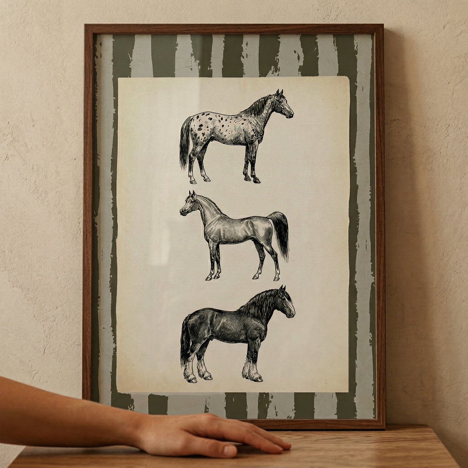A vertical wood-framed piece of art leans against a textured, neutral-toned wall. Inside the frame, an aged, off-white paper displays three detailed black and white horse sketches stacked vertically. Each horse represents a different breed: a spotted horse at the top facing right, a lean, athletic horse in the middle facing left, and a powerful draft horse at the bottom facing right. The background of the artwork features rustic, irregular vertical stripes in a dark, muted olive color.