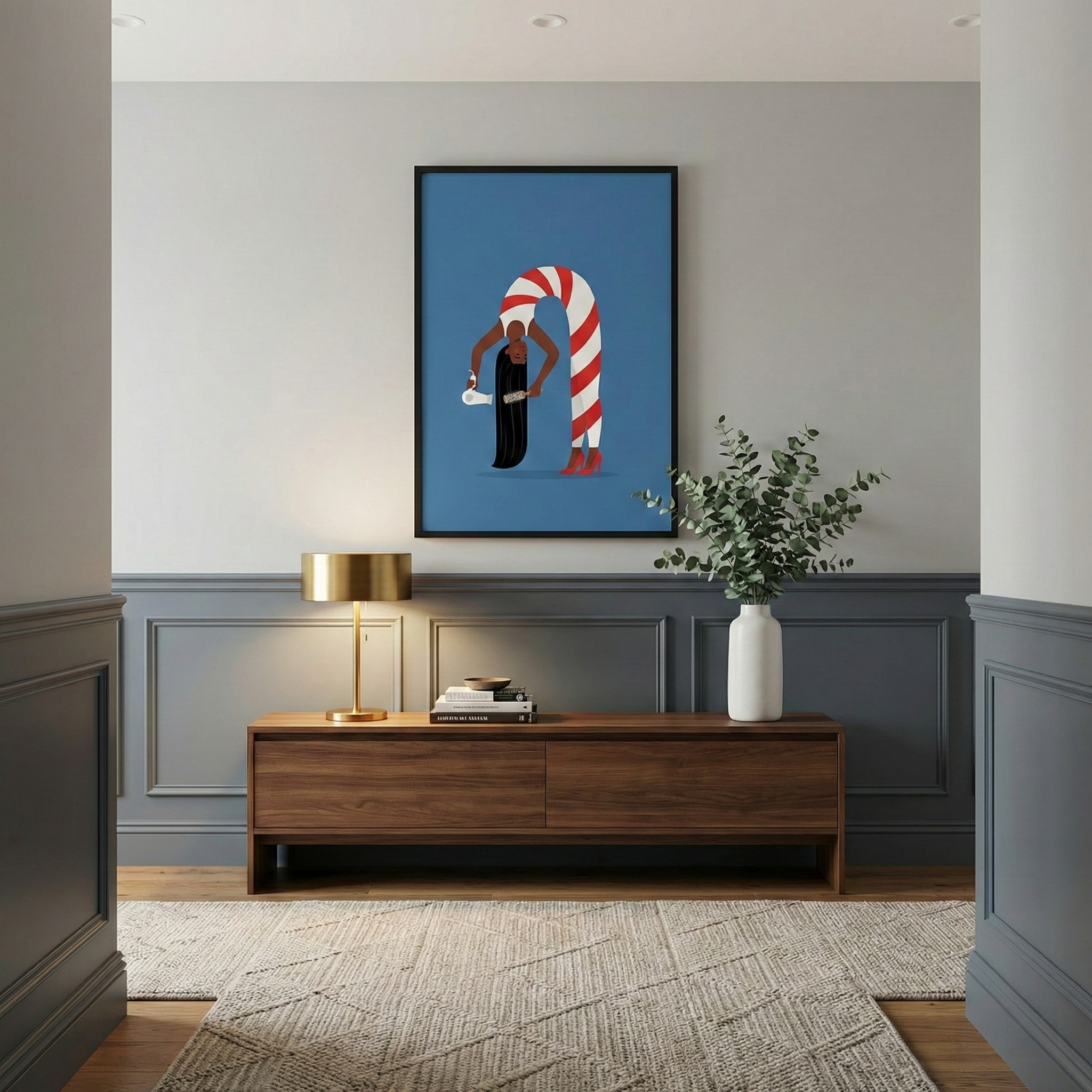 A modern interior view through a doorway. A dark wood sideboard sits against a wall with blue-gray paneling on the bottom and light gray on top. Above it hangs a framed artwork with a blue background, depicting a stylized figure with long black hair whose body forms a tall red and white striped arch. The sideboard holds a glowing gold lamp, a stack of books, and a white vase with green eucalyptus. A textured, off-white geometric rug covers the light wood floor.