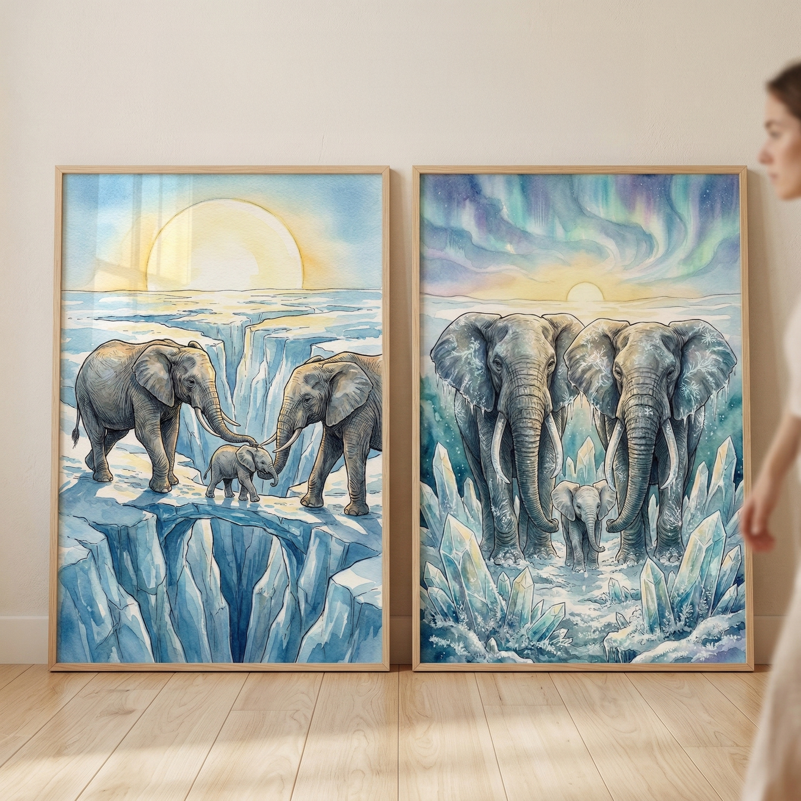 Two framed watercolor illustrations lean against a light-colored wall. The left artwork shows two adult elephants and a small calf crossing a narrow ice bridge over a deep blue crevasse under a large, glowing sun. The right artwork depicts three elephants standing among jagged ice crystals beneath a sky filled with colorful aurora borealis and a setting sun. Both pieces use soft blue, white, and gray tones. A person's arm and shoulder are partially visible on the right side of the frame.