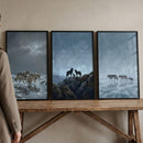 Three framed paintings show wolves moving through cold winter landscapes. One scene shows wolves walking through shallow icy water under a cloudy sky, another shows three wolves standing together on a rocky cliff with a small pup between them, and the third shows a line of wolves crossing a frozen icy surface in misty blue light. The cool muted tones create a quiet atmospheric wilderness scene.